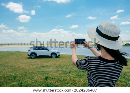 Asian woman take a picture and posed against white car in forest road with lake and bridge.