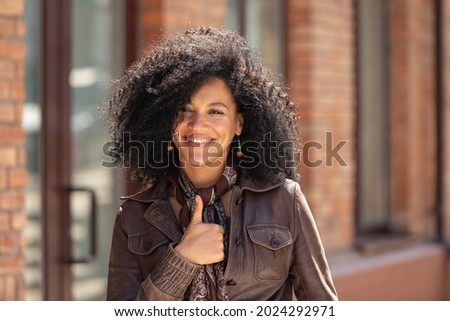 Portrait of stylish young African American woman showing thumbs up, gesture like. Brunette with curly hair in brown leather jacket posing on street against backdrop of blurred brick building. Close up