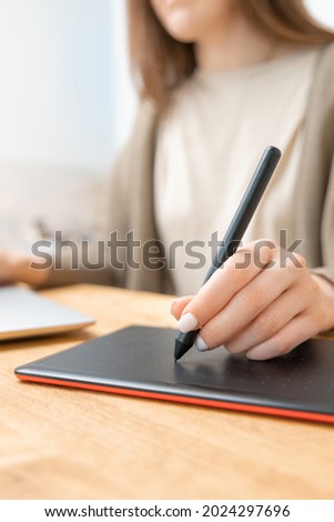 Unrecognizable woman in beige t-shirt and grey cardigan drawing or retouching photos on graphic tablet. Home office concept. Selective focus on hand with pencil, blurred background. Close up photo.