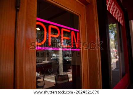 A view of a restaurant front, featuring the front door and a neon open sign in the window.