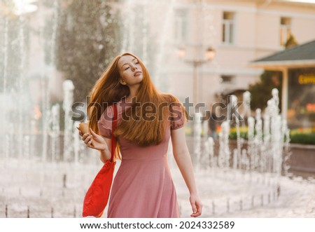 Young beautiful red-haired long-haired woman in a summer pink dress walks through the streets of a summer city.