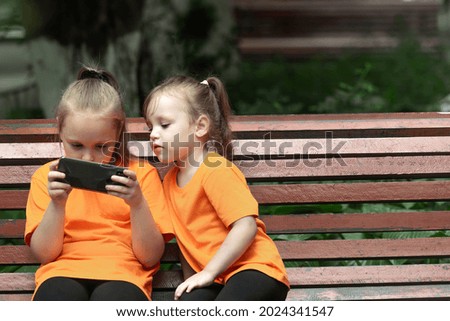 Two blonde girls of different ages in the same orange T-shirts are sitting on a bench in the open air and looking at the phone