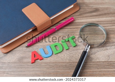 Business and audit conceptual idea.  Selective focus on AUDIT letterings made from wood.  Magnifying glass, pen, and notebook on wooden background.  Noise is visible due to the texture of the subjects