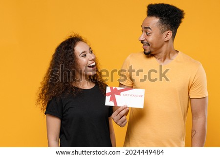 Young couple two friends together family african surprised smiling happy man woman in black t-shirt holding gift voucher flyer mock up look to each other isolated on yellow background studio portrait