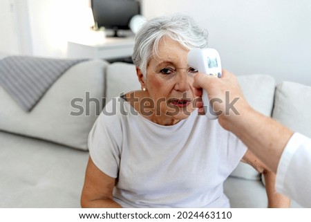 Photo of a young male doctor testing senior woman for temperature with an infrared thermometer at home. Healthcare worker at home visit. Female doctor is checking temperature to senior woman.