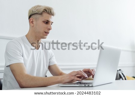 Photo of a blonde young man at the table at home talking using laptop computer.