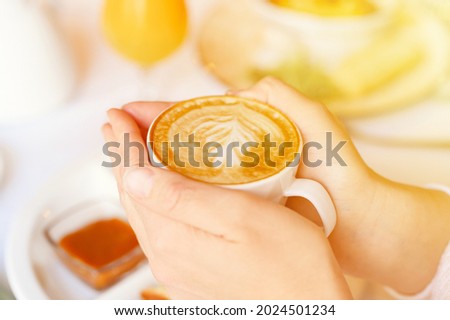 female hands holding a cup of fresh brewed coffee with beautiful leaf latte art from foam on the background of the table with a plate of food. the concept of morning breakfast and brunch. flare