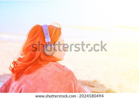 Pretty woman listening to music on the beach. Redhead girl listening to melody with listening device. Relax on a summer day at the sea. The concept of vacation. Person with headphones.