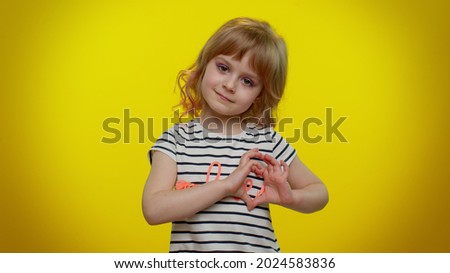 Portrait of smiling blonde child girl 5-6 years old in striped t-shirt makes heart gesture demonstrates love sign expresses good feelings and sympathy on yellow studio background. Teen kid children