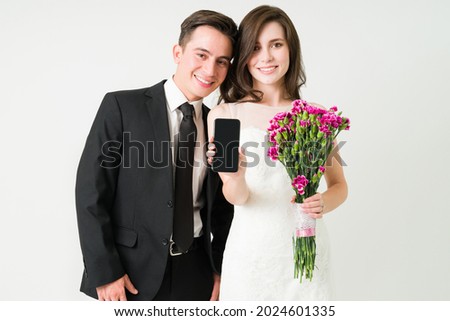 Posting our wedding picture on social media. Gorgeous bride on a wedding gown showing her smartphone screen next to the groom