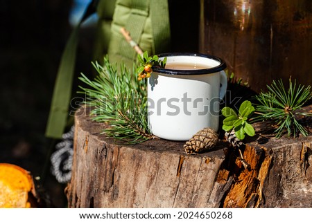White campfire enamel mug with hot herbal tea on wooden stump. Bowler pot on background, cones, forest elements as decor. Concept of lunch break during hiking, trekking, active tourism, camping