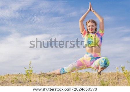 Young beautiful girl in bright multicolored tracksuit performs physical exercises to stretch muscles of legs outdoors on background of blue summer sky