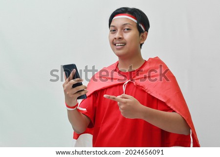Portrait of attractive Asian man in t-shirt with red white ribbon on head with flag on his shoulder as a cloak, pointing and holding mobile phone.  Isolated image on gray background
