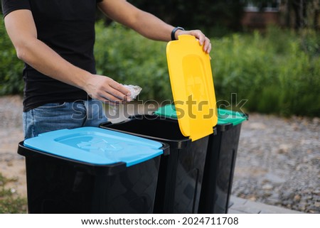 Man throwing plastic bag into recycling bin on back yard. Close up of humen hand hold rubbish. Different colours on plastic bins. Green, blue and yellow