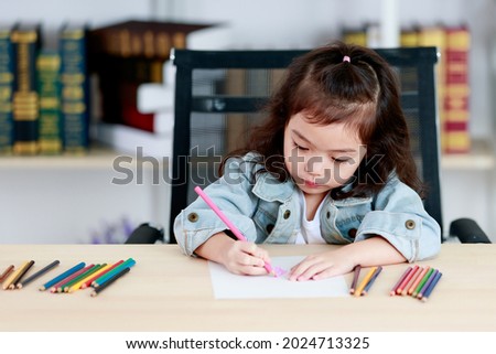 Cute little Asian girl concentrate to create picture drawing for school homework by pink pencil as art education while sitting on working desk of colored painting tools in reading room at home