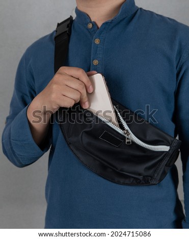 Closeup studio shot of male model in blue long sleeve shirt putting smartphone into front zipper pocket of trendy urban small black crossbody strap casual fanny pack bag in front gray background. Royalty-Free Stock Photo #2024715086