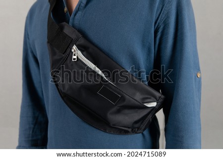 Closeup studio shot of male model in blue long sleeve shirt hanging trendy urban small black crossbody strap casual fanny pack bag in front gray background. Royalty-Free Stock Photo #2024715089
