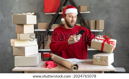 Caucasian business owner working with product packaging and smartphone wearing a Sanat Claus hat and red sweater. Scans the barcode on the box and prepares the purchase for shipment. Christmas sale