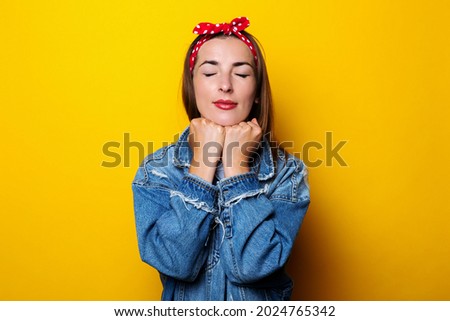 Young woman with closed eyes in a hair band, in a denim jacket holds her hands under the edge on a yellow background