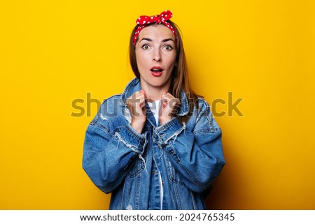 Surprised shocked young woman in hair band, in a denim jacket on a yellow background