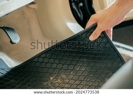 Close-up of a hand holding a dirty black rubber car floor mat with accelerator and brake inside the cabin before dusting, vacuuming, dry cleaning, washing the car. Royalty-Free Stock Photo #2024773538