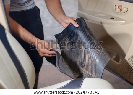 Close-up of a hand holding a dirty black rubber car floor mat with accelerator and brake inside the cabin before dusting, vacuuming, dry cleaning, washing the car. Royalty-Free Stock Photo #2024773541