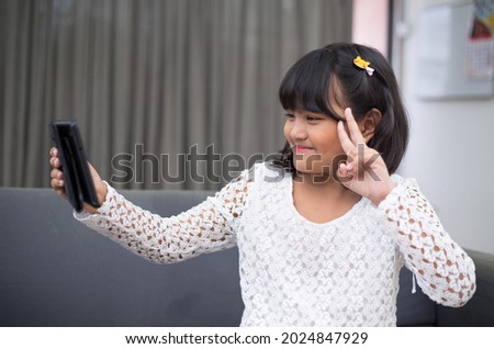 Cute and adorable little girl dressed in white, taking selfie smiling. Asian child expressing happiness as she takes selfie with tablet at home. 