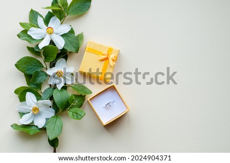 A white gold ring with a precious stone in a yellow open box with a bow lies on a beige background next to a branch with green leaves and white flowers with a yellow core with a place for text Royalty-Free Stock Photo #2024904371