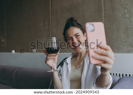 Young woman in casual clothes sit on soft grey sofa indoors apartment hold glass drink red wine do selfie shot mobile phone post photo social network Rest weekend leisure quarantine stay home concept