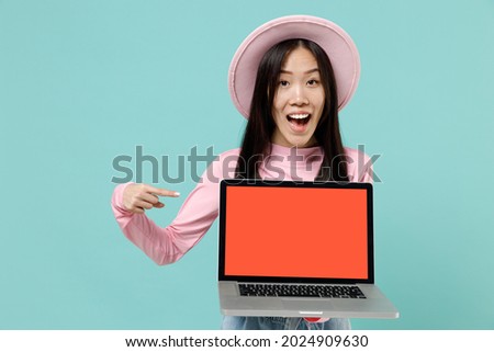 Amazed fun young brunette asian woman 20s wears pink clothes hold use work on laptop pc computer with blank screen workspace area point on it isolated on pastel blue color background studio portrait.