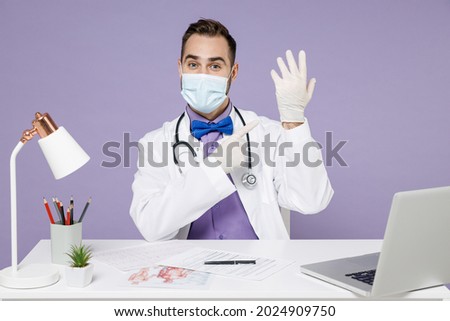 Male surgeon doctor man in white medical suit face mask sit at desk in clinic office point index finger on latex rubber gloves from coronavirus covid-19 isolated on violet background studio portrait