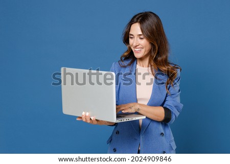 Smiling cheerful funny beautiful attractive young brunette woman 20s wearing basic casual jacket standing working on laptop pc computer isolated on bright blue colour background studio portrait Royalty-Free Stock Photo #2024909846
