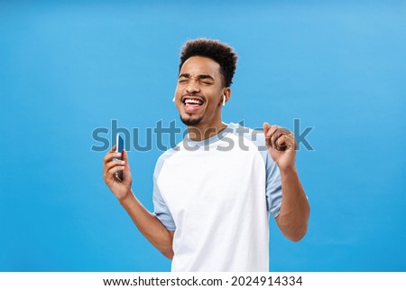 Portrait of pleased and carefree handsome stylish urban african american male in casual t-shirt dancing singing along favorite song listening music satisfied in wireless earphones holding smartphone Royalty-Free Stock Photo #2024914334