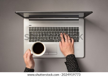 First person top view photo of woman's hands typing on laptop keyboard and holding cup of coffee on isolated grey background