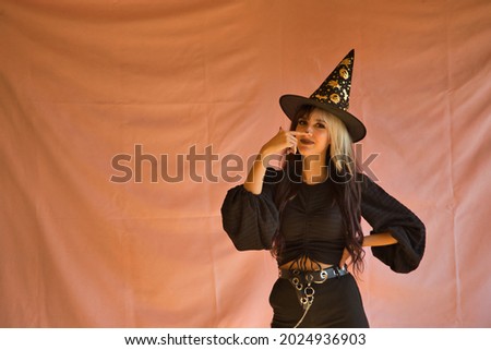 Young Hispanic woman dressed as a witch touching her nose with a finger on pink background for halloween.