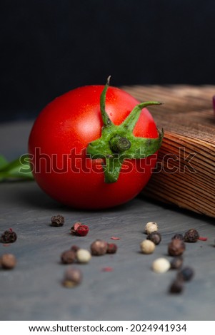 a fresh ripe tomato on a dark gray concrete table and a wooden board in the background. pepper peas scattered on the table. photo for the menu or poster. close-up. an ingredient for cooking. side view