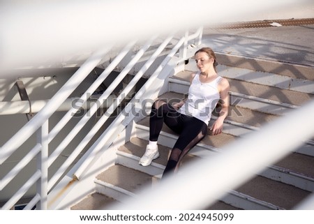 Young woman is resting while sitting on the steps of the stairs of the bridge after jogging. Beautiful background. Training in the city.