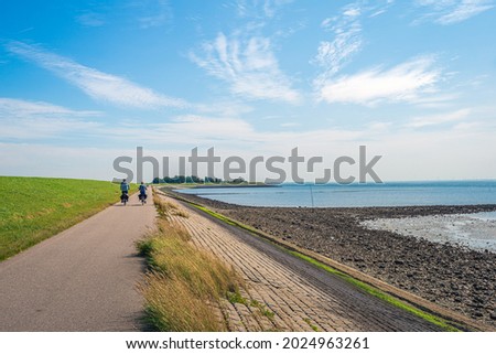 A man and a woman are cycling on a path at the bottom of a Dutch dike and along the Oosterschelde. The dike is reinforced with stones and partly covered with green algae. It is summertime now. Royalty-Free Stock Photo #2024963261