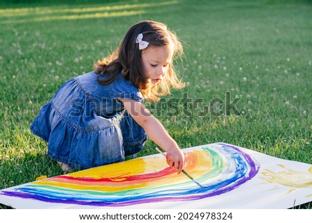 Little girl 2-4 years old paints rainbow and sun on large sheet of paper, sitting on green lawn