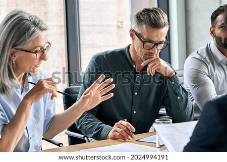 Serious mature caucasian male ceo executive manager with male business partner discussing corporation merge plan at board room table. Professional coworkers leaders doing paperwork in modern office.