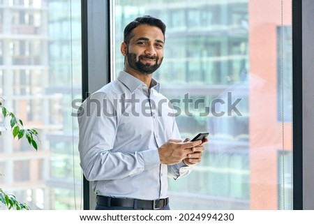 Smiling indian ceo businessman holding using cell phone mobile apps standing in office near panoramic window looking at camera. Digital tech apps and solutions for business corporate development. Royalty-Free Stock Photo #2024994230