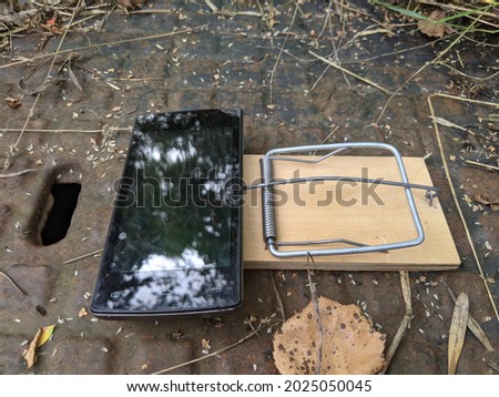 The phone is in a mousetrap. A broken black smartphone is lying on a wooden mousetrap against a background of rusty iron.