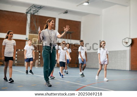 Happy physical education teacher talking to her students during a class at elementary school gym.  Royalty-Free Stock Photo #2025069218