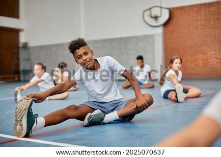 Happy black elementary student stretching his leg while warming up during physical education class at school gym and looking at camera.  Royalty-Free Stock Photo #2025070637