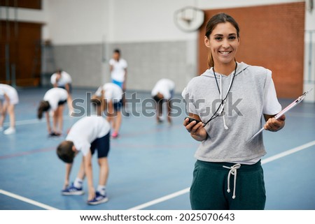 Young happy coach using stopwatch during PE class at school gym and looking at camera. Her students are exercising in the background. Royalty-Free Stock Photo #2025070643