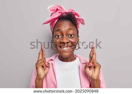 Pleasant looking black Afro American woman makes wish keeps fingers crossed anticipates positive results wears pink jacket and scarf tied over head isolated over grey background. High hopes.