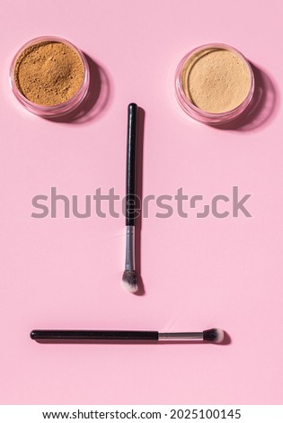 Funny face with make-up brushes and mineral powder on pink background with copy space, top view. Cosmetics humor and beauty concept.