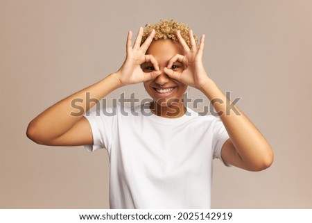 Portrait of young joyful, carefree African teenager making glasses of fingers, looking at camera with vivid smile, isolated in pale pink background with blank copy space for your advertisement