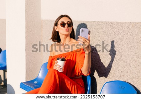 Stylish woman in orange clothes at sunset at cycle track stadium with cup of coffee and mobile phone take photo selfie Royalty-Free Stock Photo #2025153707