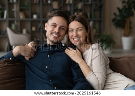 Attractive young millennial couple relax on cozy sofa at home hugging look at camera. Bank loan and mortgage, newlyweds portrait photo session, romantic relationships and love, happy marriage concept